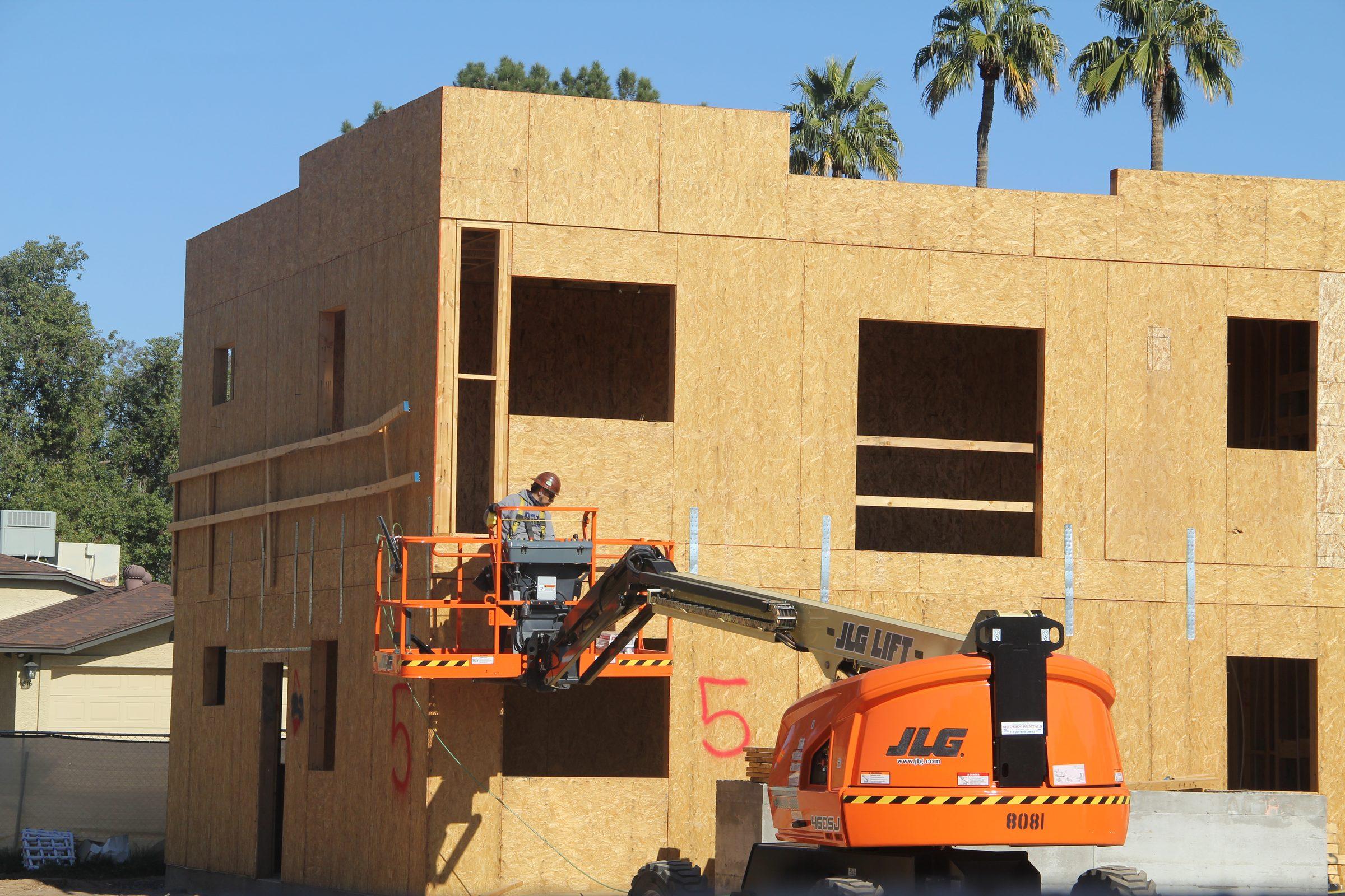 A multi-storey wood-frame building under construction, with an orange boom lift and palm trees