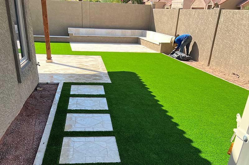 A backyard with new artificial turf and square stepping stones along a block wall